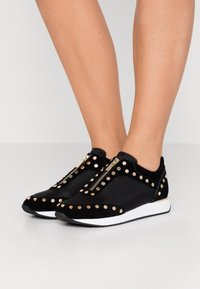 Svarta sneakers i mocka och mesh har guldnitar och dragkedjestängning, med en vit vadderad sula och rundad tådesign.