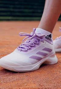 Sneaker Adidas bianchi e lavanda indossate con calzini Adidas bianchi, posizionati su una superficie di campo da tennis in terra battuta.