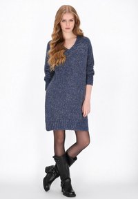 Sötétkék kötött ruha, V-nyakkal, hosszú ujjakkal és bordázott szegéllyel. Fekete térdazonos csizmával viselve. Puha tapintású, laza stílusú.