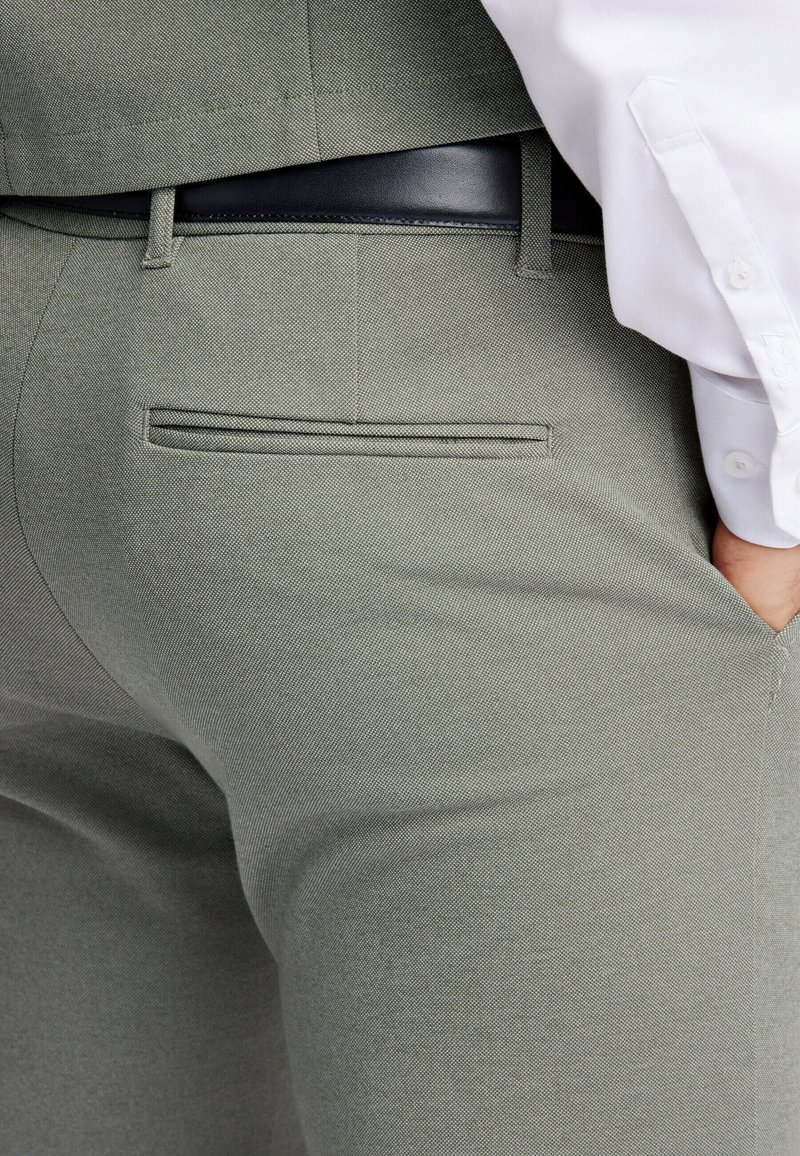 Hellgrüne, strukturierte Hosen mit schlanker Passform, versehen mit einer einzelnen Gesäßtasche und einem schwarzen Gürtel. Der Stoff hat ein dezentes Muster.
