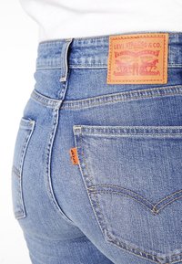 Vaqueros de mezclilla azul con una cintura alta, que presentan un parche de cuero marrón, detalles en tab naranja y bolsillos traseros con costuras en contraste.