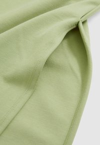 Ljusgrön tyg med en mjuk textur, som har synliga sömmar och draperade veck, vilket visar på dess lätta och ventilerande kvalitet.