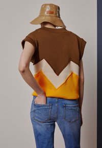Hnědý, béžový a žlutý pletený top s vzorem ve tvaru chevron. Spárován se světle modrými džíny; model má ruce v kapsách. Lesklý tanový klobouk.