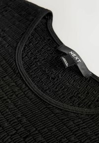 Haut noir texturé avec un col rond. Présente un design smocké pour une élasticité du tissu améliorée. Étiquette visible à l'arrière.