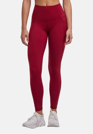 Kobieta nosząca ciemnoczerwone legginsy z wysokim stanem i bocznymi kieszeniami oraz białe buty sportowe, stojąca na jednolitym tle.