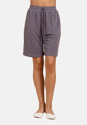 Persona che indossa pantaloncini grigi larghi fino al ginocchio con vita a coulisse e scarpe bianche basse, in piedi contro uno sfondo chiaro uniforme.