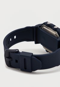 Σκούρο μπλε ψηφιακό ρολόι χειρός με ορθογώνιο καντράν και ρυθμιζόμενο λουράκι από σιλικόνη, το οποίο είναι απλωμένο επίπεδα σε λευκή επιφάνεια.