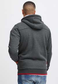 Grijze hoodie met een gestructureerde afwerking, voorzien van een rode accent bij de zoom en subtiele stikdetails op de achterkant.