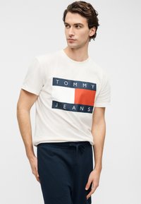 Λευκό βαμβακερό μπλουζάκι με λογότυπο σε σχήμα ορθογωνίου σε ναυτικό μπλε και κόκκινο, με το κείμενο "TOMMY JEANS". Το μοντέλο φοράει ναυτικό μπλε παντελόνια με κορδόνι στη μέση.