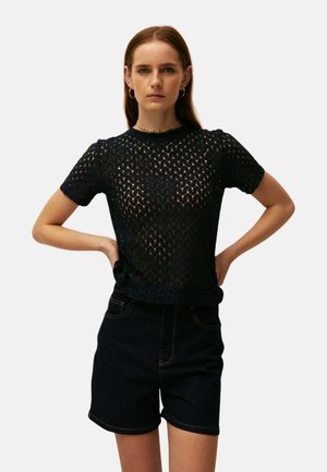 Mujer con top negro de encaje de manga corta y pantalones cortos negros de talle alto, de pie con las manos en las caderas sobre un fondo liso.