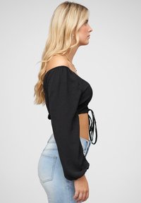 Fekete, vállról lelógó, puffos ujjú crop top, texturált anyagból, deréknál megkötős részlettel, magas derekú világoskék farmernadrággal párosítva.