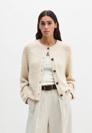 Frau mit langen dunklen Haaren, die eine beige gestrickte Strickjacke über einem weißen Oberteil, beige hoch taillierte Hosen und einen schwarzen Gürtel mit goldener Schnalle trägt.