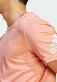 Camiseta deportiva de manga corta en rosa claro con un patrón texturizado. Presenta un cuello redondo y un logo impreso en blanco en la manga.