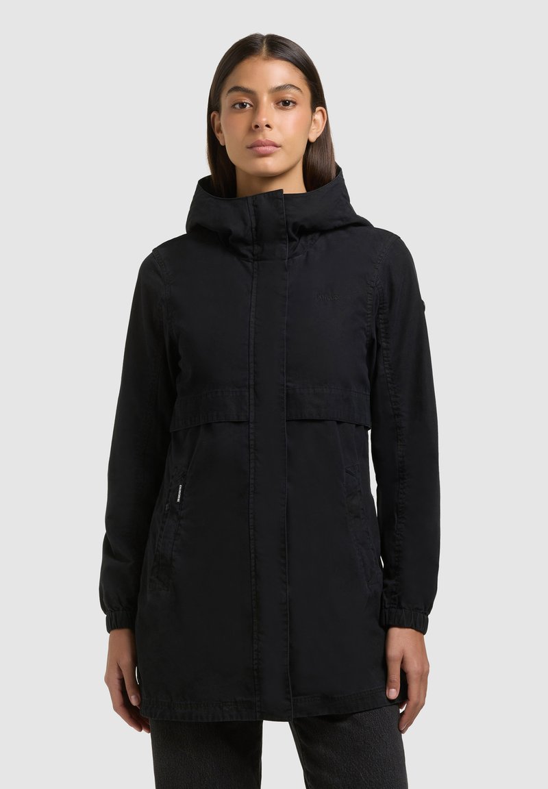 Frau mit glatten dunklen Haaren, die eine lange schwarze Kapuzenjacke mit verstecktem Reißverschluss vorne und Seitentaschen trägt und vor einem grauen Hintergrund steht.