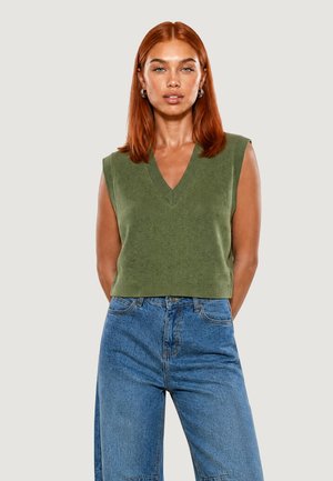 Donna dai capelli rossi che indossa un gilet verde senza maniche a scollo a V e jeans blu a vita alta, in piedi davanti a uno sfondo chiaro uniforme.