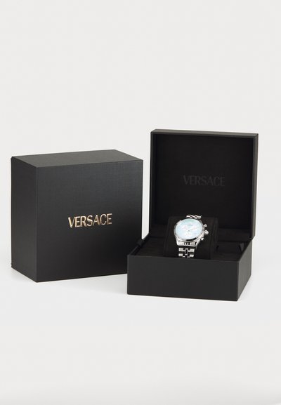 Versace-klocka i en svart låda med guldtext. Klockan har en blå urtavla, silvermetallband och en polerad finish. Lådan är texturerad.