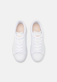 Fehér bőrsneaker, sima textúrával, lapos fehér fűzőkkel, világos barack árnyalatú akcentussal a sarkán és logóval a nyelvén.