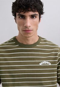 T-shirt a rayas verde oliva con líneas horizontales blancas y cuello redondo; presenta un logotipo bordado de "scalpers" en el pecho.