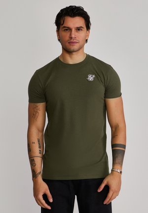 Uomo con capelli scuri indossa una maglietta verde oliva con un piccolo logo bianco sul petto, pantaloni neri e ha diversi tatuaggi sulle braccia.