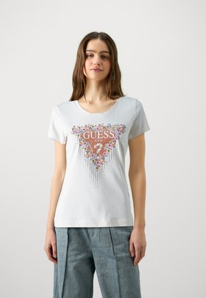 T-shirt en coton blanc avec un graphisme floral multicolore et le nom de la marque "GUESS" dans un design triangulaire, accompagné d'accents métalliques en dessous.