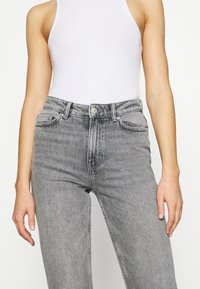 Mujer vestida con jeans gris de cintura alta y una blusa sin mangas blanca, de pie con los brazos relajados contra un fondo sencillo.