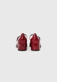 Chaussures rouges en cuir verni à talon bloc avec texture crocodile, brides à la cheville et fermetures à boucle, vues de dos sur fond gris.