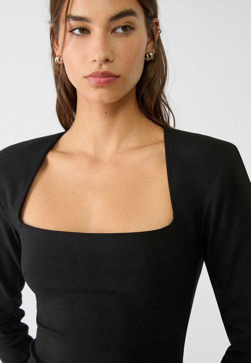 Schwarzes, tailliertes Oberteil mit quadratischem Ausschnitt und langen Ärmeln. Glatte Stofftextur, strukturierte Form und minimalistischer Stil.