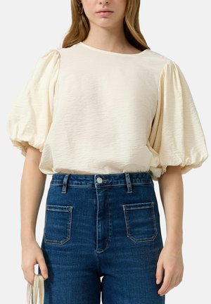 Kobieta nosząca kremową bluzkę z bufkami, wpuszczoną w ciemnoniebieskie spodnie jeansowe z wysokim stanem, trzyma kremową plecioną torbę w lewej ręce.