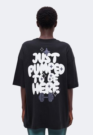 Άτομο που φοράει μαύρο υπερμεγέθες t-shirt με λευκό κείμενο «Just pumped to be here» και γραφικό με βάρη στην πλάτη.