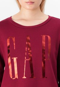 Close-up of a person wearing a maroon top with shiny copper letters "GAP" printed on the front.