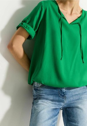 Persoon draagt een losse groene top met korte mouwen en strikjes, gecombineerd met lichtblauwe spijkerbroek, staand met één arm gebogen achter de rug.