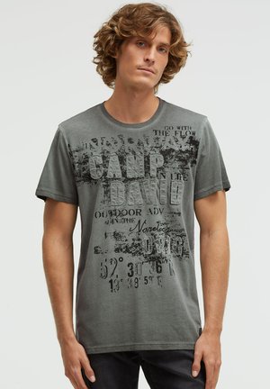 Junger Mann mit lockigem Haar, der ein graues T-Shirt mit verblasstem schwarzem Text und Koordinaten trägt, vor einem schlichten hellen Hintergrund steht.