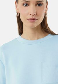 Lichtblauwe sweatshirt met een ronde hals en korte mouwen, voorzien van een klein borstzakje en een gladde textuur.