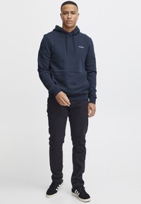 Hanorac bleumarin cu siret, buzunar kangaroo și țesătură texturată. Asortat cu pantaloni negri și adidași negri cu dungi albe.