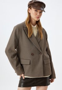 Blazer doppiopetto marrone a quadri con collo a rever, due tasche frontali e bottoni marroni. Indossato sopra un maglione crema.