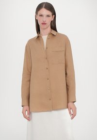 Brown button-up shirt with a chest pocket and embroidered initials, made of textured fabric, paired with a white, shiny skirt.