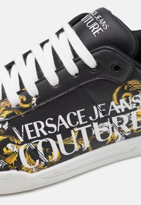Svart syntetisk sneakers med vita snören, med framträdande vit text "VERSACE JEANS COUTURE" och guldfärgade utsmyckade designaccenter.