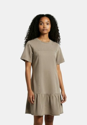 Donna con capelli ricci che indossa un abito taupe a maniche corte, lungo fino al ginocchio, con orlo arricciato e semplici dettagli di cucitura.
