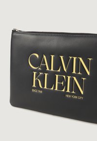 Sort læderclutch med sølvlynlås. Har fed gul tekst: "CALVIN KLEIN," "SINCE 1968," og "NEW YORK CITY." Glat tekstur.