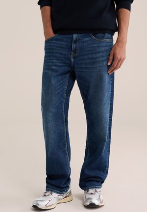 Homme portant un jean droit bleu foncé, un pull noir et des chaussures de course blanches, debout avec une main dans la poche.
