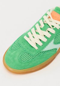 Grønn sneaker med en kombinasjon av semsket skinn og mesh-materialer, tykke kremfargede snørebånd og en gummisåle for godt grep. Merkbare lyseblå detaljer.