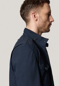 Granatowa, lekka kurtka z wysokim kołnierzem, bocznymi kieszeniami i dodatkami w postaci zatrzasków. Nieco teksturowana tkanina i usztywniony krój.