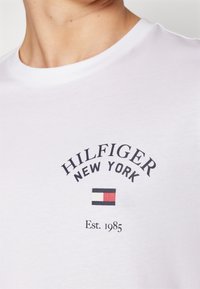 Weißes Baumwoll-T-Shirt mit Rundhalsausschnitt. Verfügt über den Schriftzug "HILFIGER NEW YORK" und ein rechteckiges Logo mit roten und blauen Akzenten. Jahr "Est. 1985" ist enthalten.