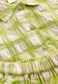 Robe en plaid vert et beige avec un col, des boutons en bois et un détail de taille froncée. Tissu en coton au toucher doux.