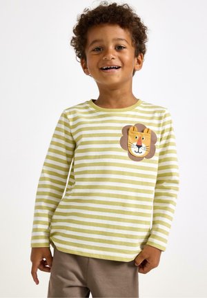 Bambino sorridente con capelli ricci che indossa una maglietta a maniche lunghe a righe verde e bianca con una toppa a forma di leone e pantaloni marroni su uno sfondo bianco.
