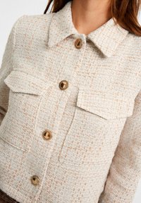 Geweven textuurjack in crème met subtiele perzikaccenten. Beschikt over een kraag, twee voorzakken en gouden knopen.
