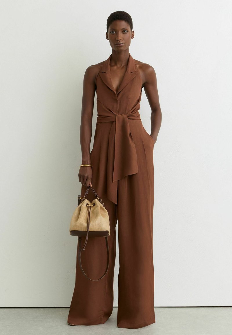 Kvinde iført brun ærmeløs jumpsuit med vide ben og knyttet talje, holder en beige skuldertaske, stående mod en hvid væg.