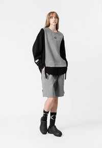 Sudadera oversized gris y negra con un gran bolsillo, pantalones cortos negros y calcetines con estampado. El calzado son zuecos negros texturizados.