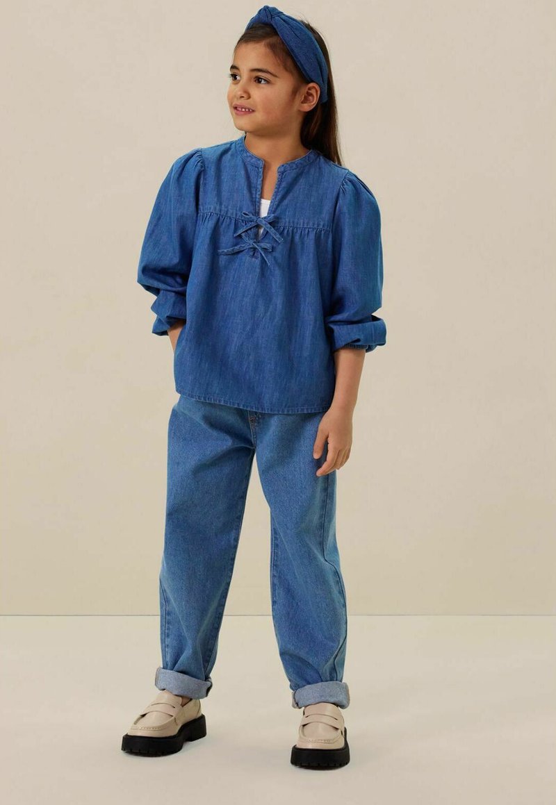 Haut en denim bleu avec col et détail de nouage, associé à un jean bleu clair retroussé et des chaussures beige à enfiler, sur un fond neutre.