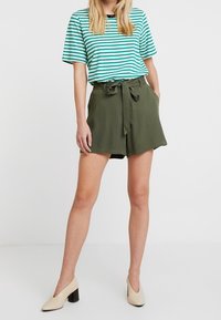 Camicia a strisce turchese e bianca a maniche corte abbinata a pantaloni corti verde oliva a vita alta con cintura in tessuto. Il modello indossa scarpe con tacco beige.